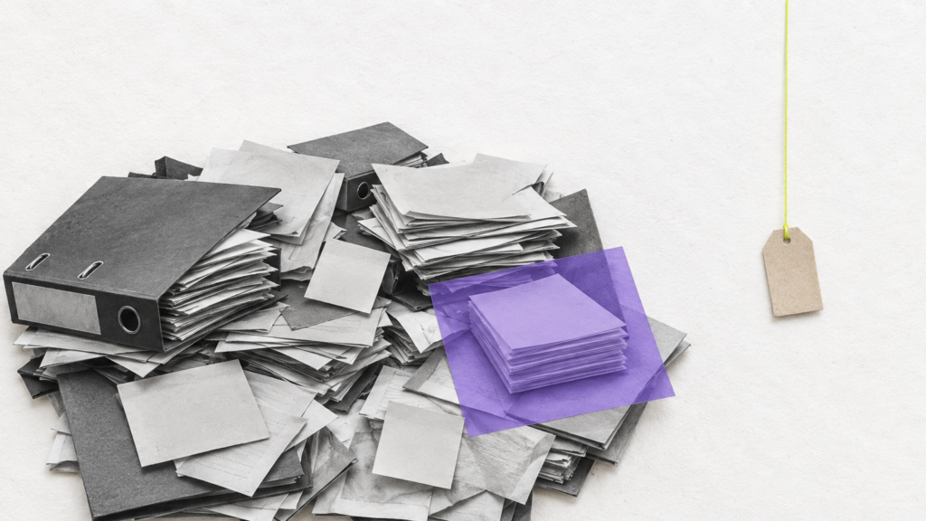 Una gran pila desordenada de carpetas y papeles en blanco y negro ocupa la izquierda; un pequeño montón queda ordenado bajo papel morado. A la derecha cuelga una etiqueta de precio vacía y diminuta, mientras el trabajo de organizar el proceso conserva el peso visual.
