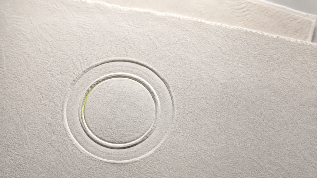 Macro de un sello en relieve seco impreso sobre papel grueso de color crema; el relieve se distingue solo por su sombra, sin tinta ni emblema legible, con un filo de luz verde en el borde interior del círculo
