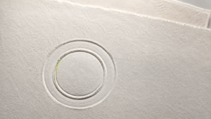 Macro de un sello en relieve seco impreso sobre papel grueso de color crema; el relieve se distingue solo por su sombra, sin tinta ni emblema legible, con un filo de luz verde en el borde interior del círculo